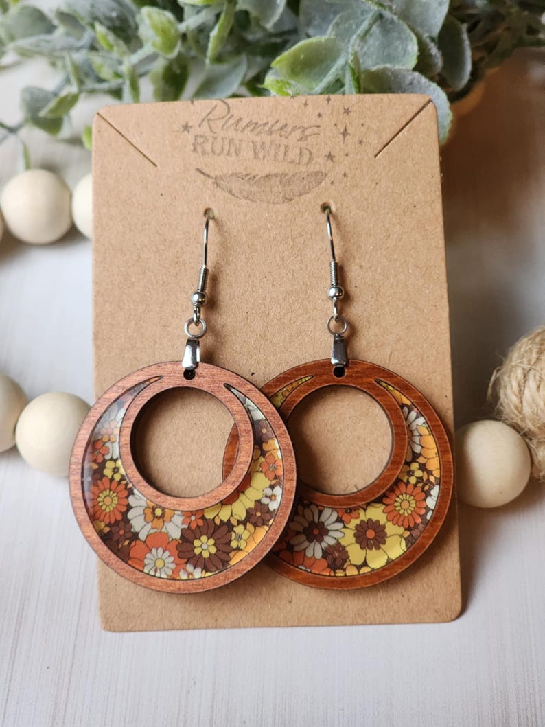 May include: Wooden hoop earrings with a retro floral design in shades of yellow, orange, brown, and white. The earrings are attached to a brown card with the text "Rumurs Run Wild".
