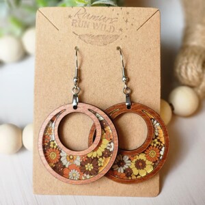 May include: Wooden hoop earrings with a retro floral design in shades of yellow, orange, brown, and white. The earrings are on a brown card with the text "Rumurs Run Wild". The earrings have silver-colored hooks.