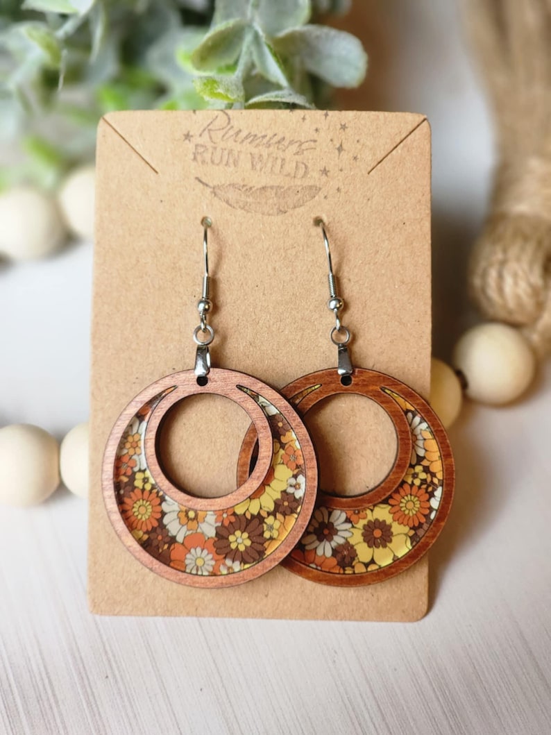 May include: Wooden hoop earrings with a retro floral design in shades of yellow, orange, brown, and white. The earrings are on a brown card with the text "Rumurs Run Wild". The earrings have silver-colored hooks.