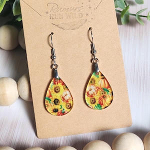 May include: Teardrop-shaped dangle earrings featuring a vibrant print of yellow sunflowers and orange pumpkins. The earrings hang from silver-toned metal hooks and are displayed on a tan card printed with the text Rumors RUN WILD.