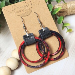Puede incluir: Pendientes colgantes con cuadrados negros de imitaci&oacute;n cuero y aros estampados en rojo y negro. Los pendientes se exhiben en una tarjeta marr&oacute;n con el texto "Rumurs Run Wild". Ganchos plateados.