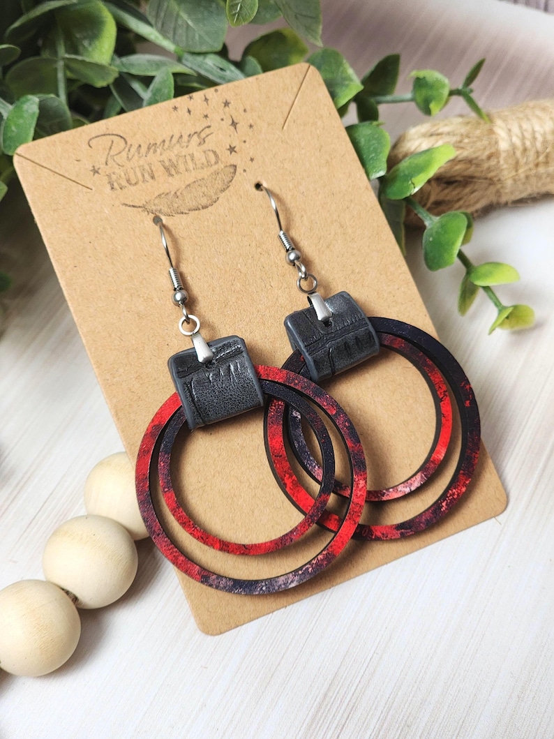 Puede incluir: Pendientes colgantes con cuadrados negros de imitaci&oacute;n cuero y aros estampados en rojo y negro. Los pendientes se exhiben en una tarjeta marr&oacute;n con el texto "Rumurs Run Wild". Ganchos plateados.