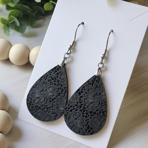 May include: A pair of black teardrop-shaped earrings with a delicate floral pattern. The earrings are made of leather and have silver hooks.