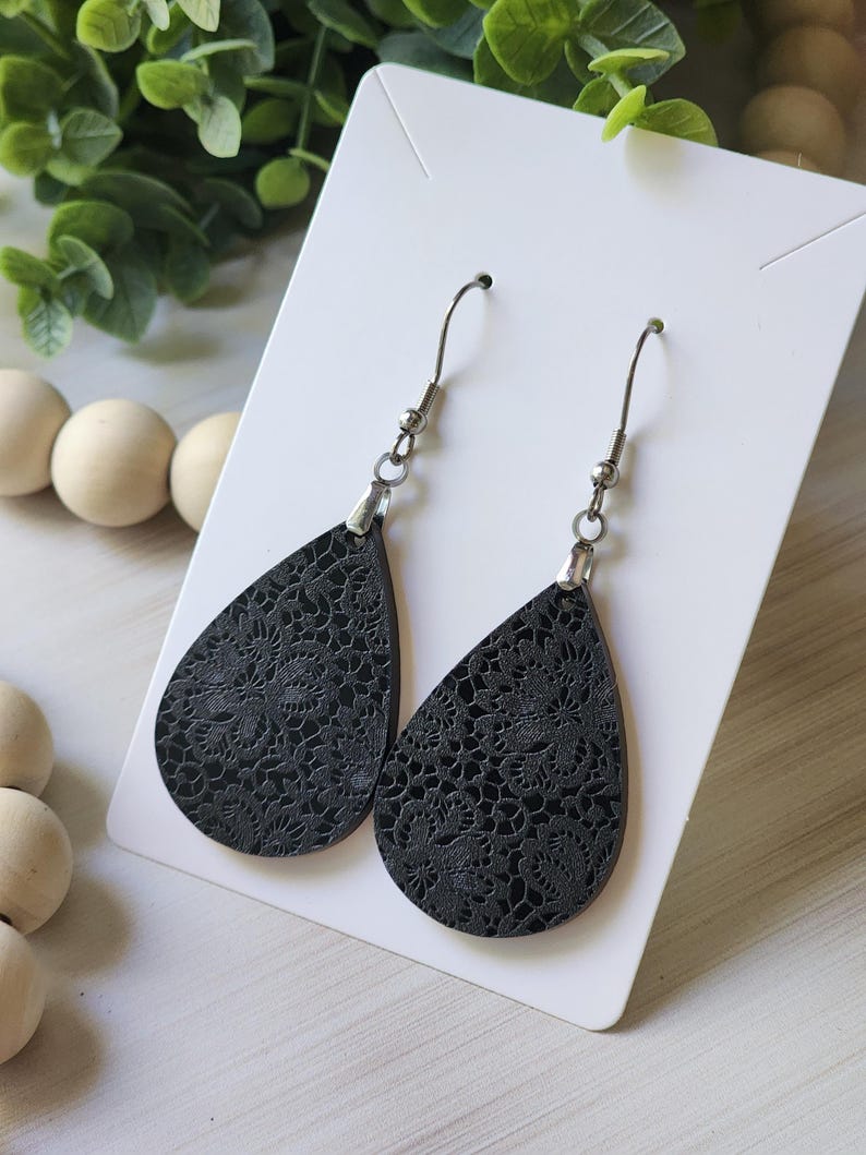 May include: A pair of black teardrop-shaped earrings with a delicate floral pattern. The earrings are made of leather and have silver hooks.