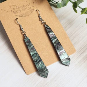 May include: A pair of dangle earrings with a marbled green, purple, and silver design. The earrings are rectangular with a pointed bottom and are suspended from silver-colored hooks. The earrings are displayed on a brown card with the text "Rumors Run Wild".