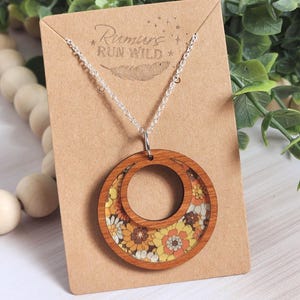 May include: A wooden pendant necklace with a silver chain. The pendant features two concentric circles, the outer one is wood, and the inner one has a floral pattern in yellow, orange, and white. The necklace is displayed on a brown card with the text "Rumurs Run Wild".
