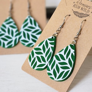 Può includere: Un paio di orecchini a goccia verde e bianco con un motivo a foglia. Gli orecchini sono su una carta marrone con il testo "Run Wild" stampato su di essa.