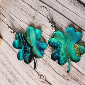 May include: A pair of silver dangle earrings with four-leaf clover charms. The clovers are made of green and blue resin with gold glitter accents.