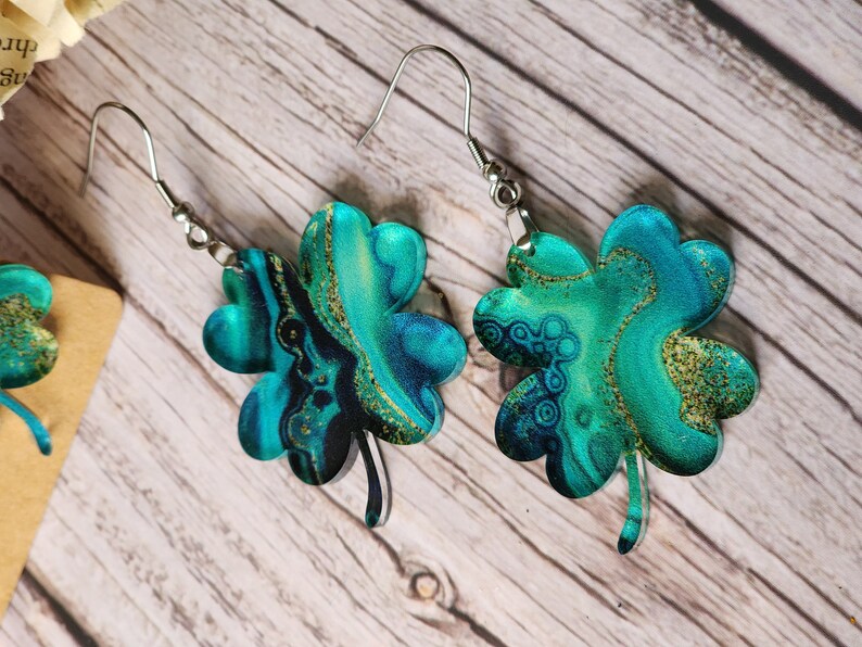 May include: A pair of silver dangle earrings with four-leaf clover charms. The clovers are made of green and blue resin with gold glitter accents.