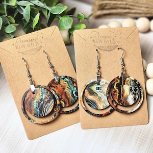 May include: Two pairs of dangle earrings with a circular design. The earrings feature a mix of colors including brown, orange, teal, and white. The earrings are displayed on brown cardboard earring cards. The text "Rumors Run Wild" is printed on the cards.