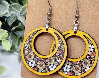 May include: A pair of wooden earrings with a floral pattern and a yellow border. The earrings are in the shape of a crescent moon and have silver hooks.