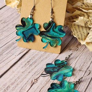 May include: A pair of green and gold four-leaf clover earrings with a marbled design. The earrings are hanging from a brown card with the text "Rumurs RUN WILD" on it.