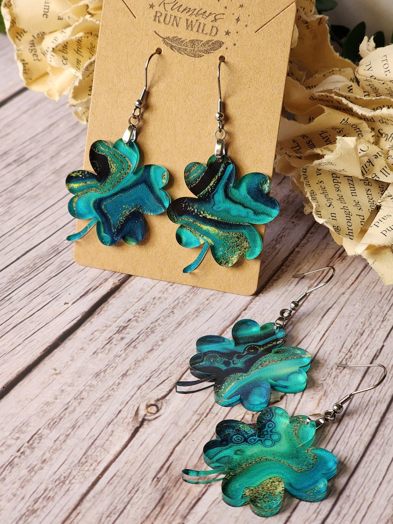 May include: A pair of green and gold four-leaf clover earrings with a marbled design. The earrings are hanging from a brown card with the text "Rumurs RUN WILD" on it.