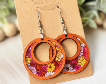 May include: A pair of wooden earrings with a floral pattern. The earrings are orange with a white and pink floral design. The earrings are in the shape of a circle with a smaller circle cut out of the center. The earrings are hanging from silver hooks.