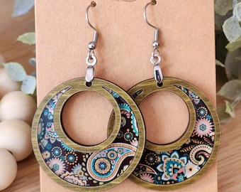 May include: A pair of wooden earrings with a paisley pattern in shades of blue, green, and black. The earrings are round and have a gold-colored metal hoop. The earrings are hanging on a card that says "Rimmers Run Wild".