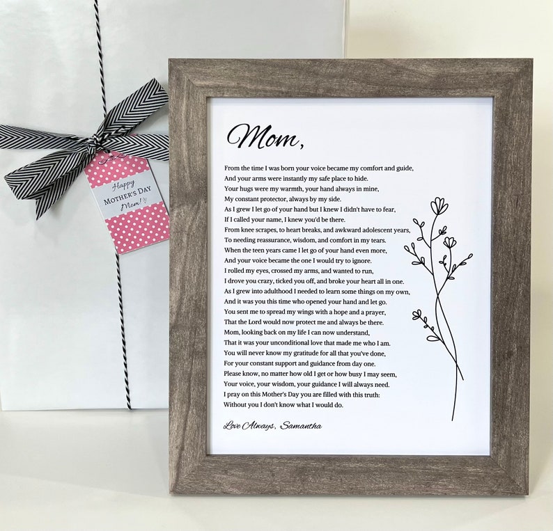 May include: A framed print with a poem titled "Mom" in black text, with a floral illustration. The frame is gray. A gift wrapped in white paper with a black and white ribbon and a pink tag that says "Happy Mother's Day Mom!" is on the left.