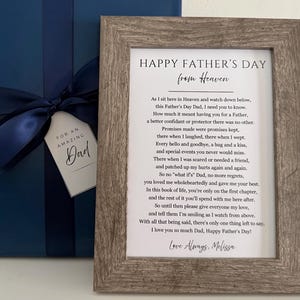 May include: A rectangular picture frame with a weathered wood finish containing a printed poem titled Happy Father's Day from Heaven. Beside it sits a dark blue gift box wrapped with a matching satin ribbon and a tag reading For An Amazing Dad.