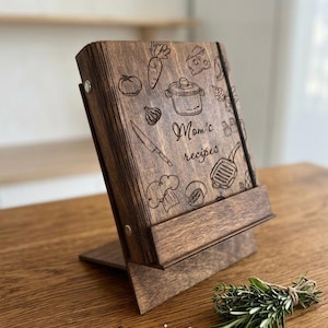 May include: A dark brown wooden recipe book stand featuring an engraved design of kitchen utensils, vegetables, and the text "Mom's recipes". The stand is angled to hold a book and is constructed from stained wood with visible grain patterns.
