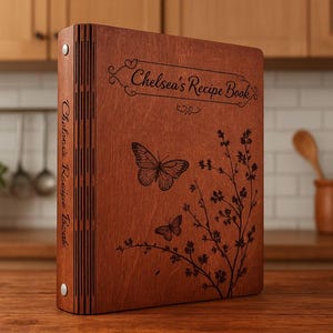 Puede incluir: Un libro de recetas de madera con el texto "Chelsea's Recipe Book" grabado en la portada. La portada presenta un diseño de mariposa y flores. El libro es marrón y tiene una encuadernación de tres anillas.