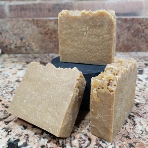 May include: Three bars of handmade soap, each with a light brown color and a slightly rough texture. The soap bars are stacked on top of each other.