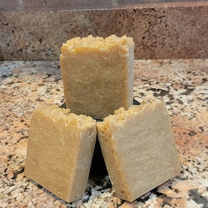 May include: Three bars of handmade soap, each with a light brown color and a rough texture. The bars are stacked on top of each other, with the top bar slightly tilted to the side.