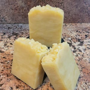 May include: Three bars of handmade soap, each a light yellow hue. The tops of the soap have a rough, uneven texture, while the sides are smooth, displaying swirls. The soap is arranged on a dark surface, set against a speckled background. Each bar is approximately 8 cm in length.