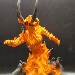 May include: A hand-painted miniature figurine of a fiery demon with black horns and a swirling orange and yellow flame body. The demon is standing on a base of gray fabric.