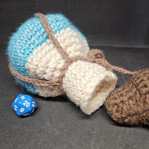 May include: A crocheted dice bag with a blue, white and brown striped design. The bag is tied with a brown cord and has a small brown crocheted heart attached to it. A blue twenty-sided die is lying next to the bag.