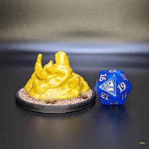 May include: A yellow, 3D printed miniature of a rocky terrain, resembling a mound of dirt or sand, on a black base with brown grass. A blue, 20-sided dice with gold numbers is in the background.