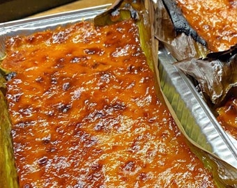 Può includere: Primo piano di un dolce al forno di colore marrone dorato, probabilmente una prelibatezza filippina chiamata "bibingka", avvolto in foglie di banana e appoggiato su una teglia d'argento.