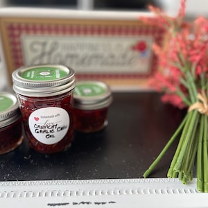 May include: Three glass jars filled with red chili oil and garlic. The jars have green lids and white labels with black handwritten text that reads "Homemade with Love Crunchy Garlic Chili Oil".