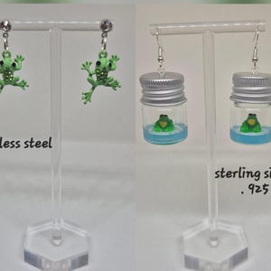 May include: Two pairs of earrings are displayed. One pair features green frog charms with rhinestone accents, labeled "stainless steel." The other pair has small frog figurines inside miniature glass jars, labeled "sterling silver .925."