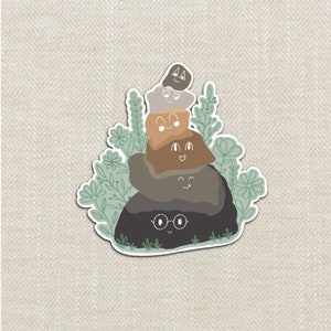 May include: A stack of four grey, brown, and black rocks with cute faces and eyes. The rocks are surrounded by green leaves and white flowers.