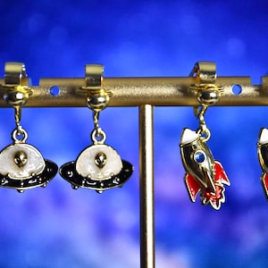 May include: A set of four gold-tone earrings with a space theme. Two earrings feature a white and black UFO with a green alien face. The other two earrings feature a gold rocket with red and blue accents.