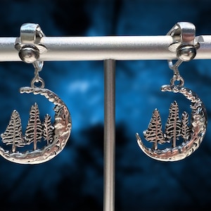 May include: Silver clip-on earrings featuring a crescent moon design with three stylized trees inside.