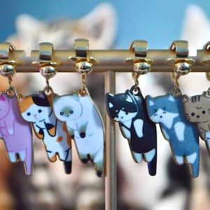 Op de afbeelding: Acht gouden oorringen met kattencharms in verschillende kleuren en patronen. De charms hangen aan een gouden standaard.