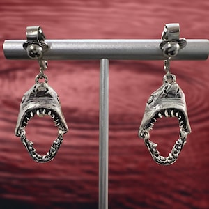 May include: A pair of silver clip-on earrings featuring a shark's open mouth with sharp teeth. The earrings are displayed on a silver stand against a red background.