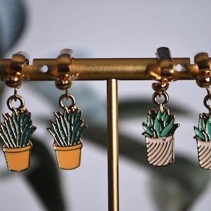 May include: A pair of gold-tone clip-on earrings with a dangling charm of a potted succulent plant. The succulent is green and the pot is yellow. The earrings are displayed on a gold stand.