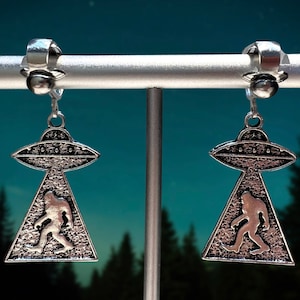 May include: A pair of silver clip-on earrings featuring a UFO with a Bigfoot silhouette inside. The earrings are on a display stand against a blue and black starry night background.