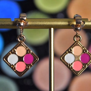 May include: A pair of gold-tone clip-on earrings with a square-shaped charm featuring four colorful circles in shades of pink, peach, white, and purple.