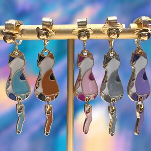 May include: Five pairs of gold cat-shaped clip-on earrings. Each cat is painted with a different color scheme: blue, brown, pink, gray, and purple.