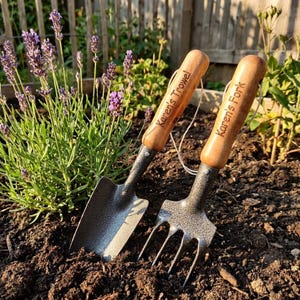 Puede incluir: Dos herramientas de jardiner&iacute;a, una paleta y un tenedor, se muestran en un parterre. Los mangos de madera de las herramientas llevan grabadas las inscripciones "Karen's Trowel" y "Karen's Fork". Las herramientas est&aacute;n plantadas en tierra oscura, con plantas de lavanda al fondo.