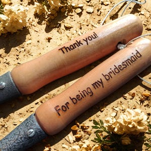 Puede incluir: Dos paletas de jard&iacute;n de madera con mangos de color marr&oacute;n claro y hojas de metal gris. Una paleta dice "Thank you!" y la otra "For being my bridesmaid". Las paletas est&aacute;n sobre mantillo marr&oacute;n con flores secas.