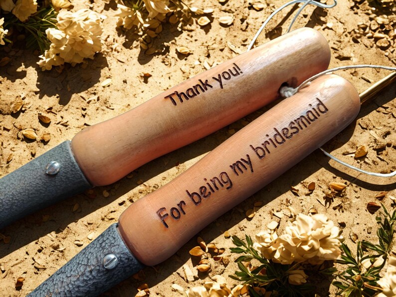 Puede incluir: Dos paletas de jard&iacute;n de madera con mangos de color marr&oacute;n claro y hojas de metal gris. Una paleta dice "Thank you!" y la otra "For being my bridesmaid". Las paletas est&aacute;n sobre mantillo marr&oacute;n con flores secas.