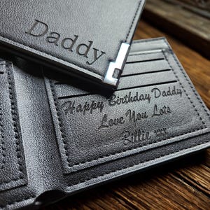 May include: A black bi-fold wallet with a silver-toned metal corner accent. The exterior is engraved with the word Daddy. The interior features a clear ID window and an engraved message reading Happy Birthday Daddy Love You Lots Billie xxx.