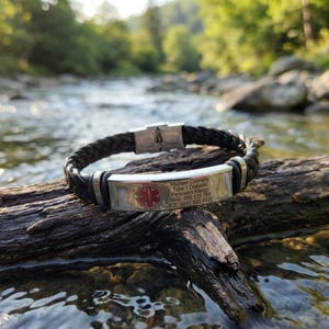 May include: Black braided leather medical ID bracelet with a stainless steel clasp and plate. The plate has a red medical symbol and text: Michael Connelly, Type 1 Diabetic, Penicillin Allergy, NHS: 456 123 789, ICE: 0845 525 4526.