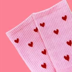 May include: A pair of light pink socks with a ribbed texture. The socks are adorned with a pattern of small, dark red hearts. The background is a solid, matching pink.