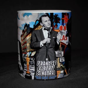 May include: A white ceramic mug featuring a black and white graphic of Frank Sinatra singing into a microphone. The design includes the text "FRANCIS ALBERT SINATRA" and "OLD BLUE EYES," along with imagery of an Oscar statue, city buildings, and neon signs.