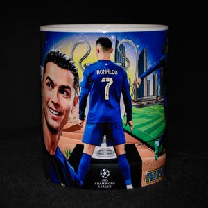 Puede incluir: Taza de cerámica blanca con un diseño que presenta a un jugador de fútbol con uniforme azul con el nombre "Ronaldo" y el número "7". La taza también incluye el logotipo de la Liga de Campeones y un fondo de paisaje urbano.