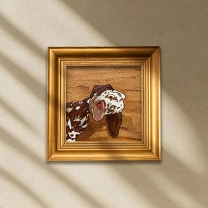 May include: A framed artwork featuring a brown and white spotted dog with its tongue out. The dog is set against a wood-grain background, and the frame is gold. The artwork is square and hangs on a beige wall.